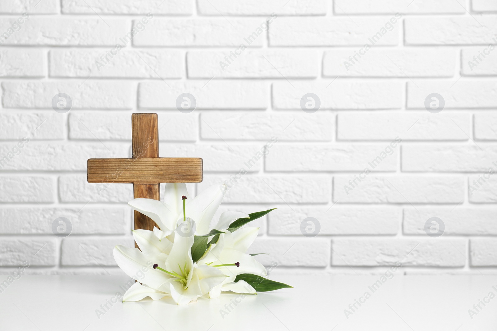 Photo of Wooden cross and blossom lilies on table against brick wall, space for text