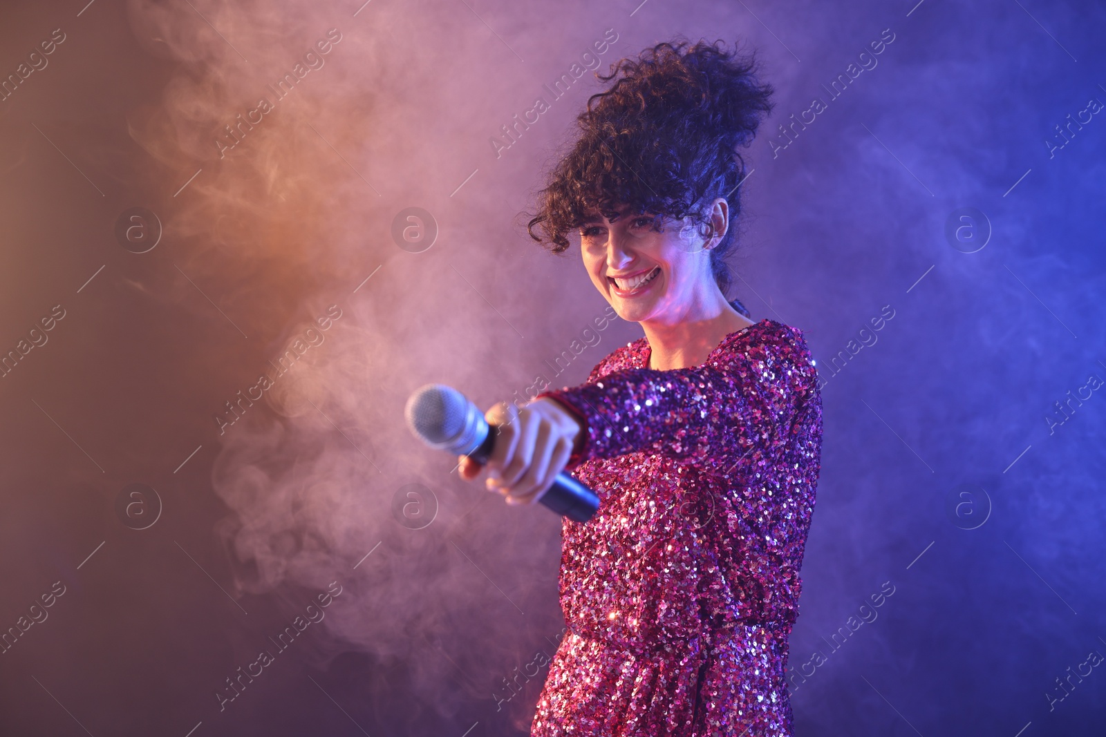 Photo of Beautiful young woman with microphone on color background in neon lights and smoke