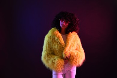 Beautiful young woman in yellow fur coat and sunglasses on color background in neon lights