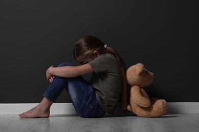 Photo of Child abuse. Upset girl with toy sitting on floor near grey wall