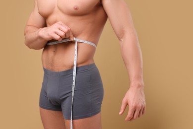 Photo of Athletic man measuring waist with tape on brown background, closeup. Weight loss concept