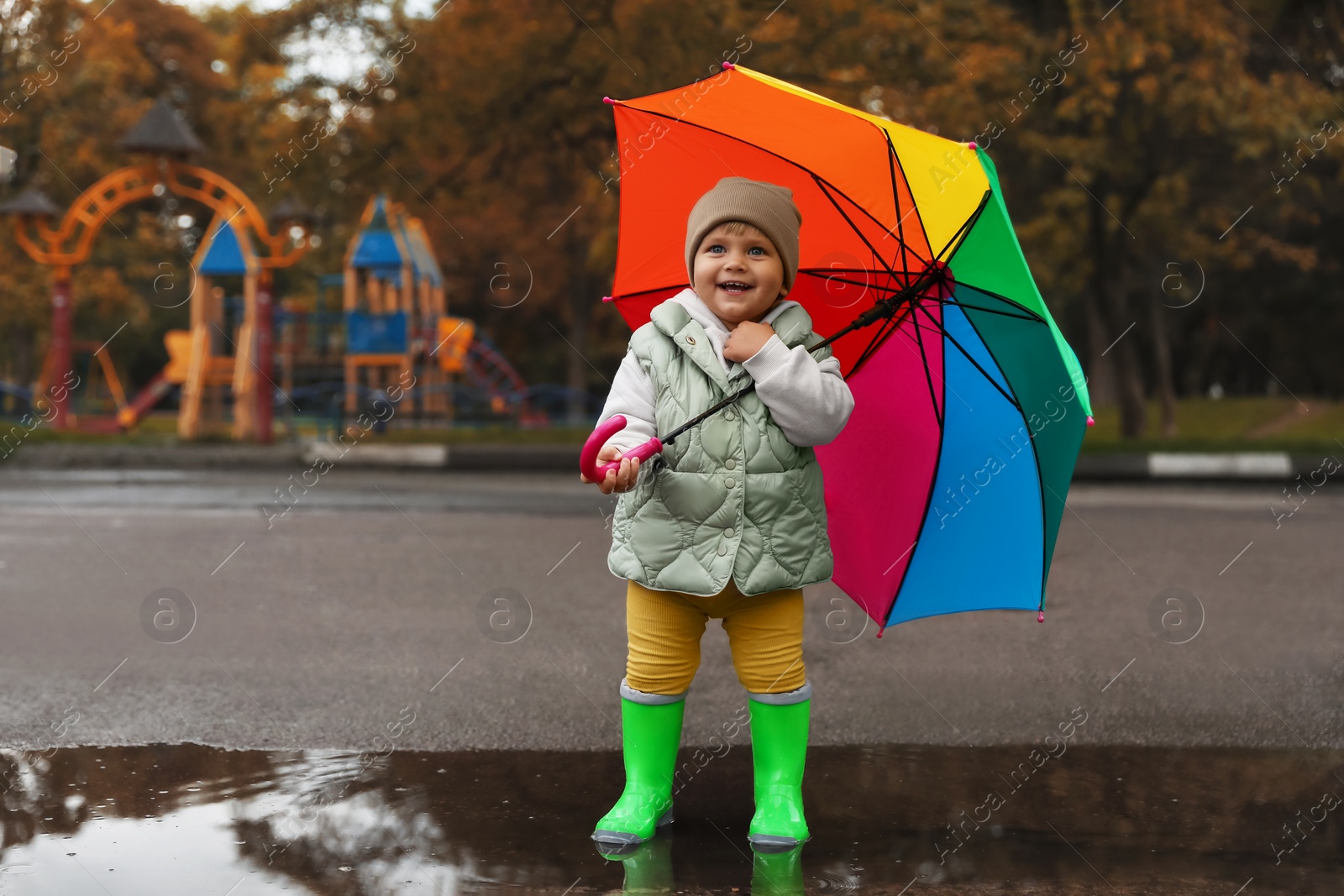 Photo of Cute little girl with colorful umbrella standing in puddle outdoors