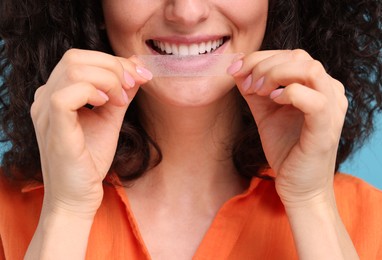 Young woman applying whitening strip on her teeth against light blue background, closeup