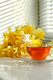 Photo of Cup of aromatic tea and beautiful yellow daffodils on windowsill. Space for text