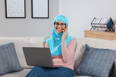Muslim woman talking on smartphone near laptop at couch in room