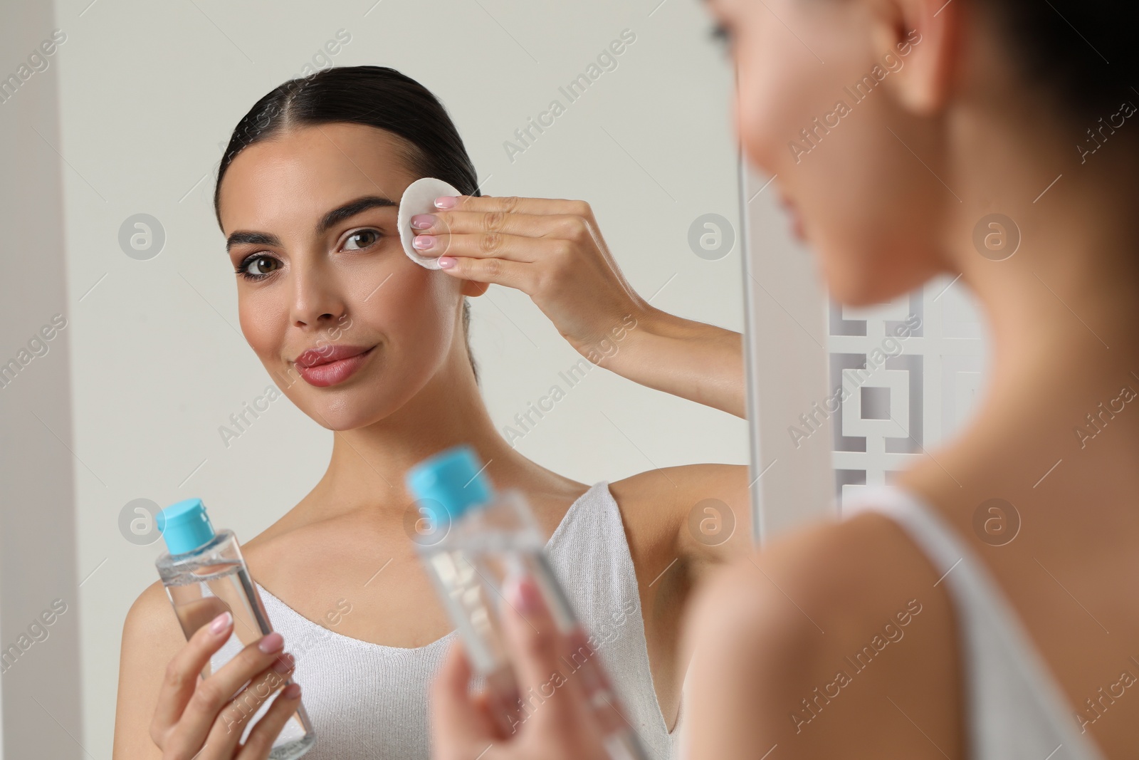 Photo of Beautiful woman removing makeup with cotton pad near mirror indoors