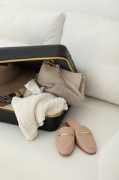 Open suitcase full of clothes, jacket and fashionable shoes on sofa