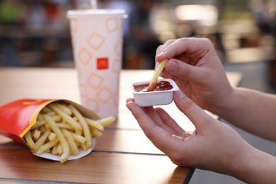Photo of Lviv, Ukraine - September 26, 2023: Woman dipping McDonald's french fry into sauce at wooden table outdoors, closeup