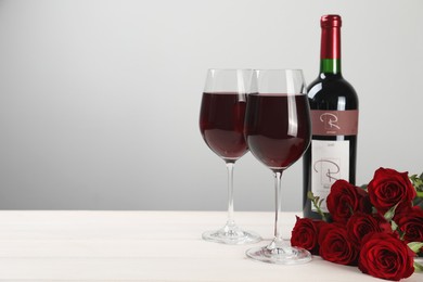 Bottle and glasses of red wine near beautiful roses on white wooden table. Space for text