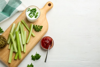 Celery sticks with different sauces and greenery on white wooden table, top view. Space for text