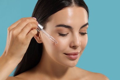 Beautiful young woman applying serum onto her face on light blue background, closeup