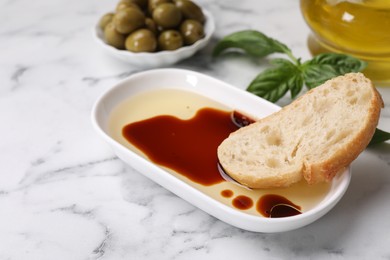 Photo of Bowl of organic balsamic vinegar with oil served with bread slice, basil and olives on white marble table. Space for text
