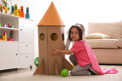 Cute African American child playing with cardboard rocket at home