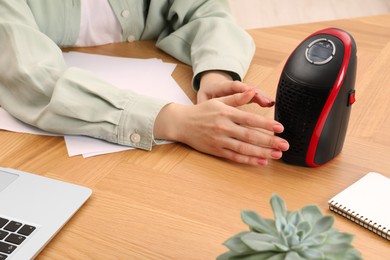 Photo of Young woman warming hands near compact electric heater at wooden table, closeup