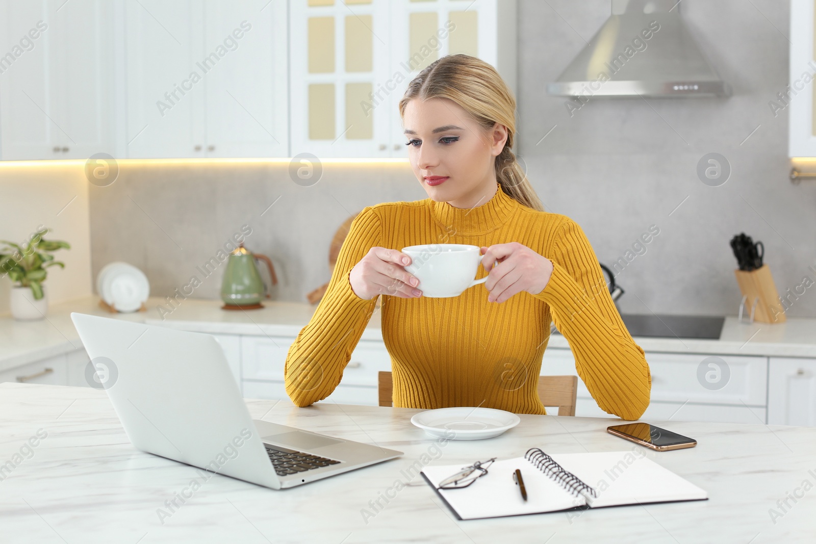 Photo of Home workplace. Woman with cup of hot drink looking at laptop at marble desk in kitchen