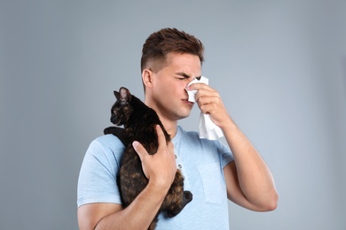 Young man with cat suffering from allergy on grey background