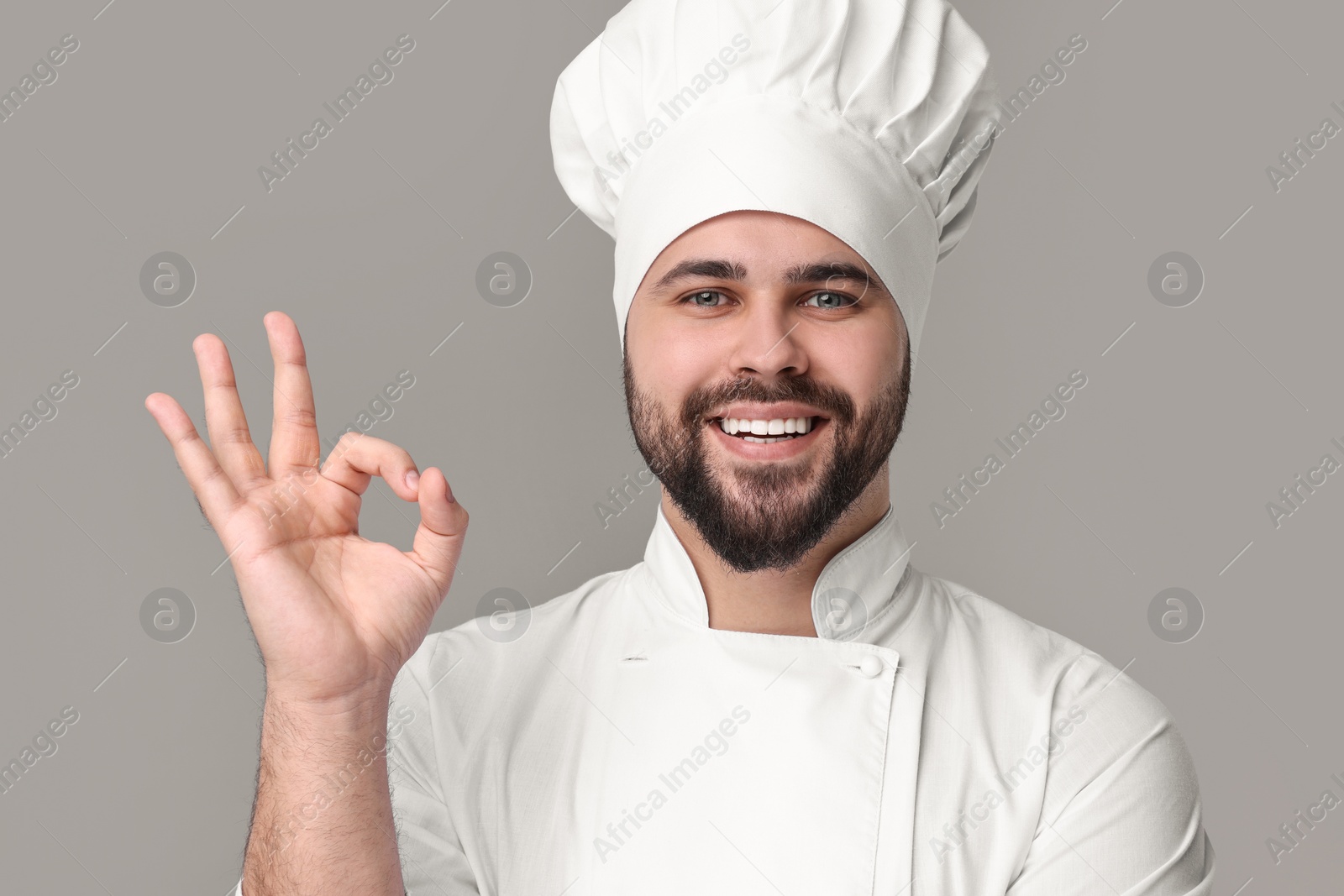 Photo of Happy young chef in uniform showing ok gesture on grey background