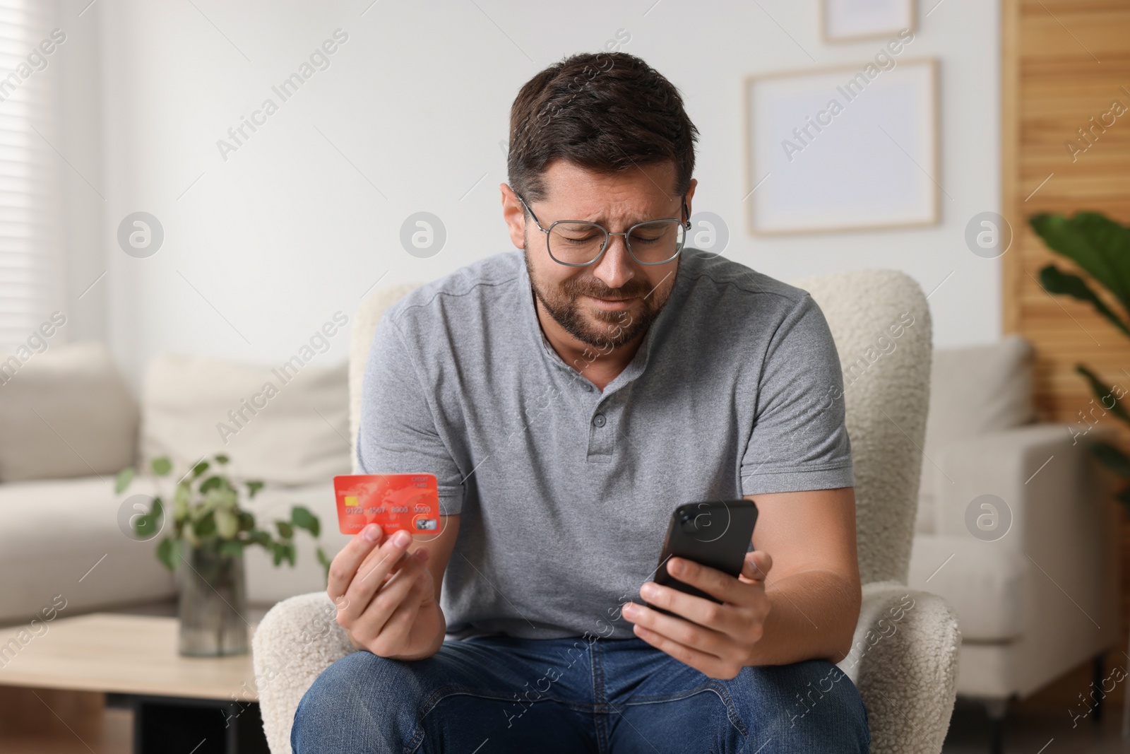 Photo of Emotional man with credit card and smartphone in armchair at home. Be careful - fraud