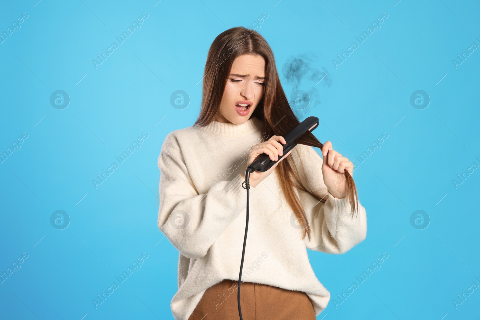 Image of Stressed young woman with flattening iron on light blue background. Hair damage