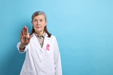 Photo of Mammologist with pink ribbon showing stop gesture on light blue background, space for text. Breast cancer awareness