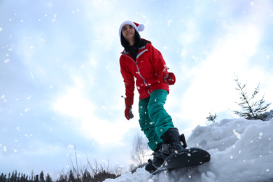 Photo of Young snowboarder wearing Santa hat on snowy hill, low angle view. Winter vacation