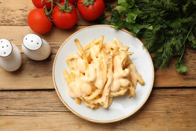 Photo of Delicious French fries with cheese sauce, tomatoes, dill and parsley on wooden table, flat lay