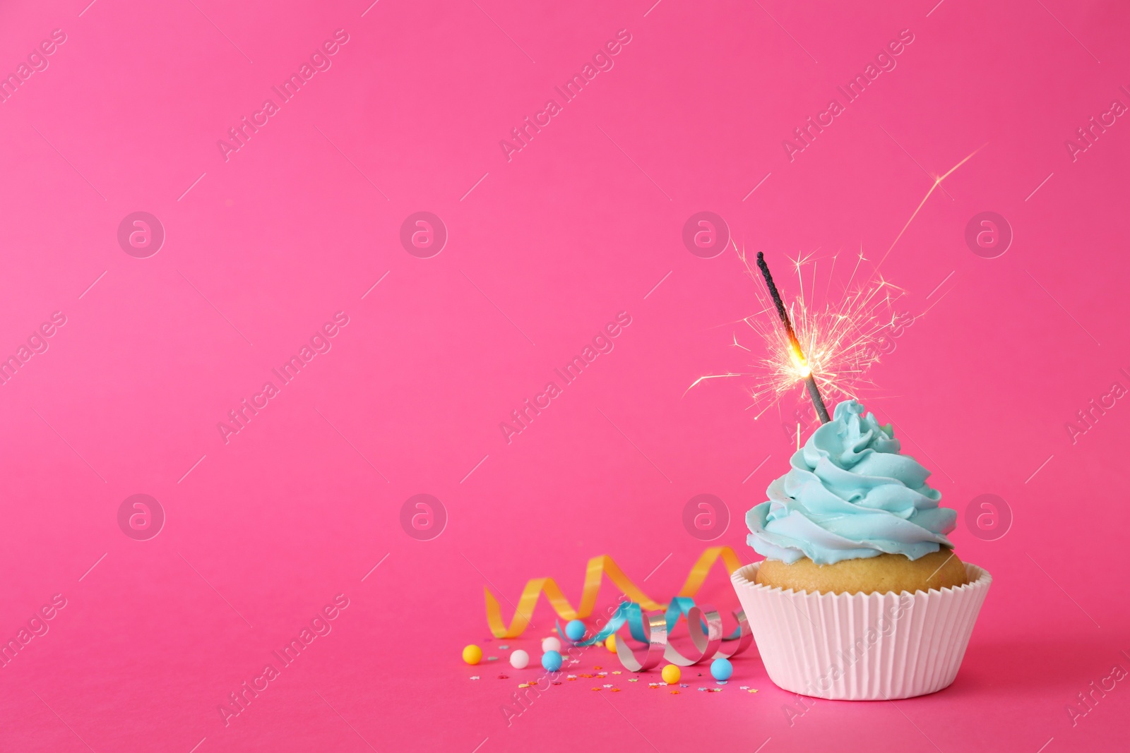 Photo of Birthday cupcake with burning sparkler and streamers on pink background. Space for text