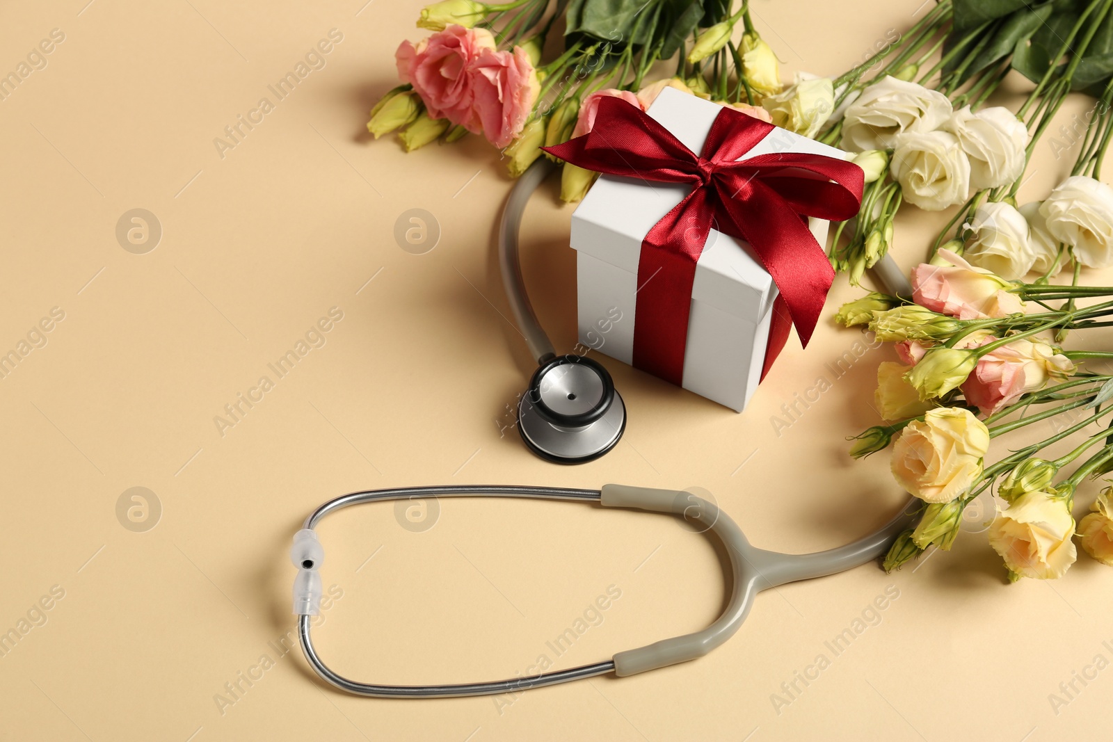 Photo of Stethoscope, gift box and flowers on beige background. Happy Doctor's Day