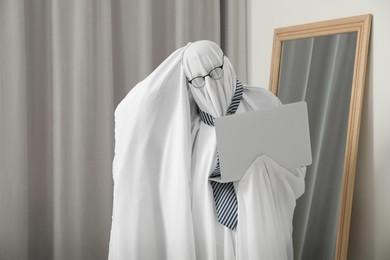 Photo of Overworked ghost. Man in white sheet using laptop at home