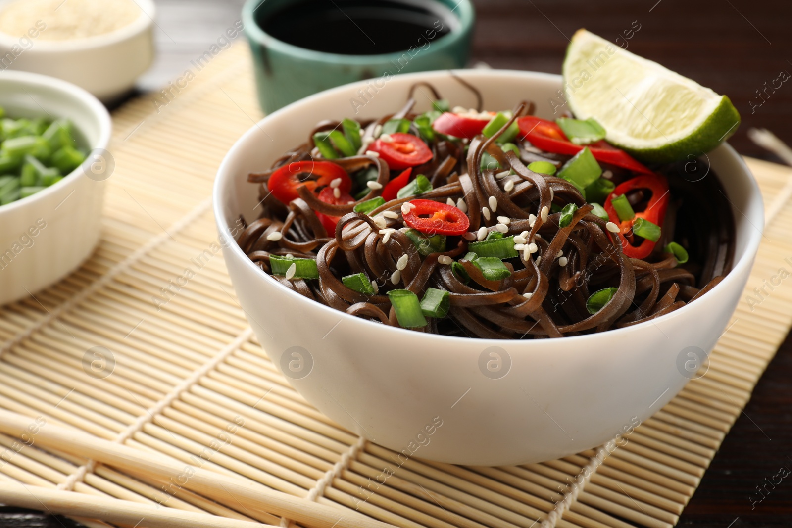 Photo of Tasty buckwheat noodles (soba) with chili pepper, onion, sesame and lime in bowl on table, closeup