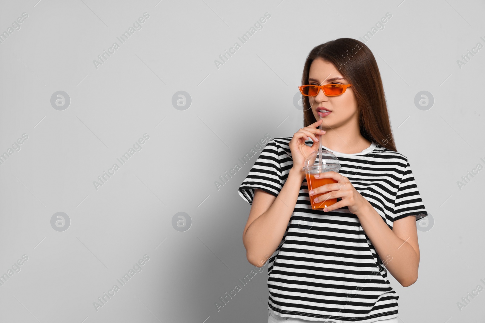 Photo of Beautiful young woman drinking juice from plastic cup on light grey background. Space for text