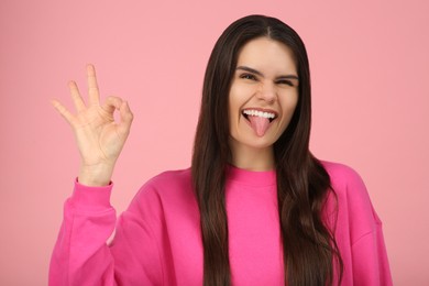 Happy young woman showing her tongue and making ok gesture on pink background