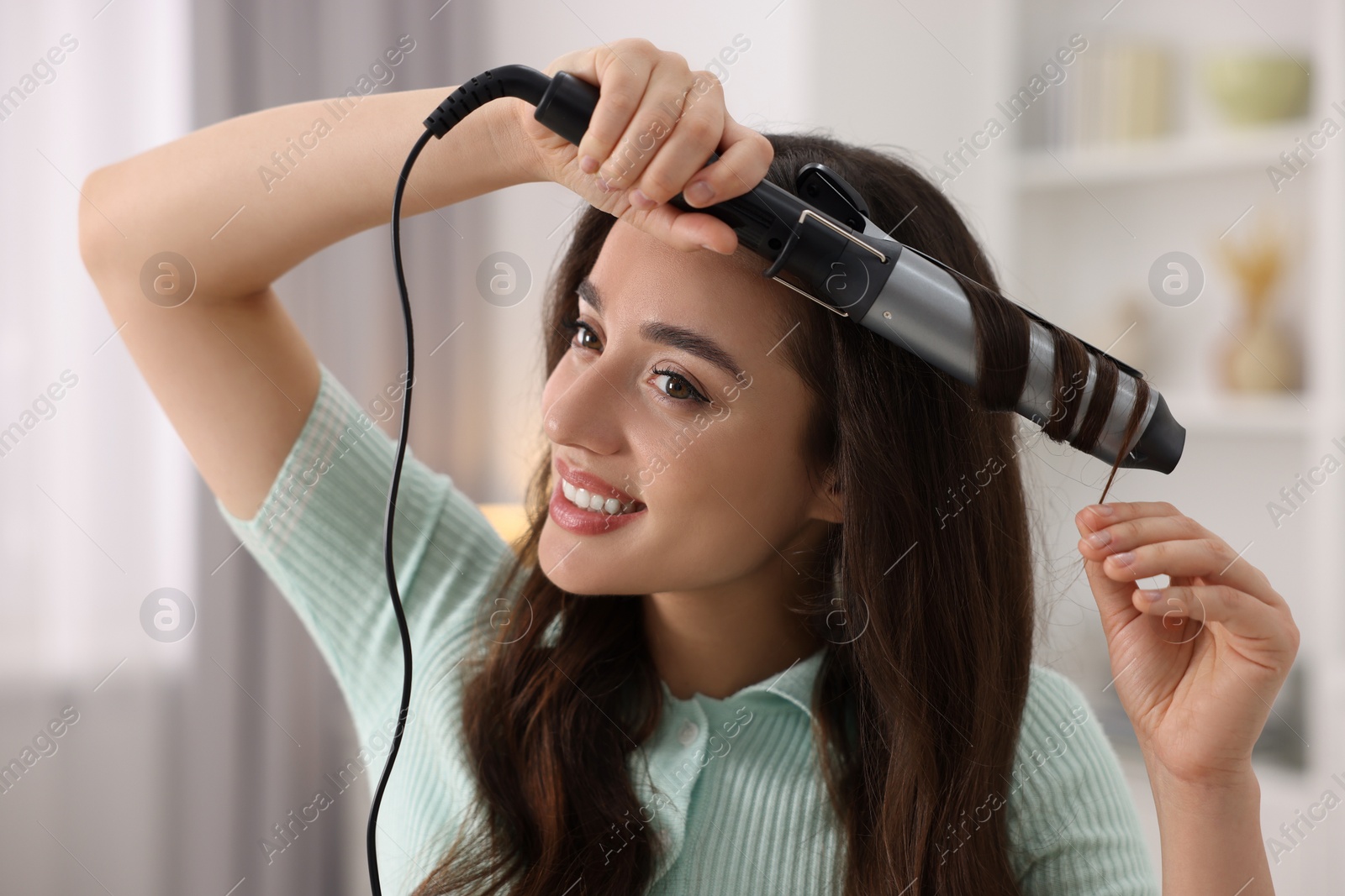 Photo of Smiling woman using curling hair iron indoors