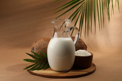 Photo of Glass jug of delicious vegan milk, coconuts, flakes and palm leaf on brown background