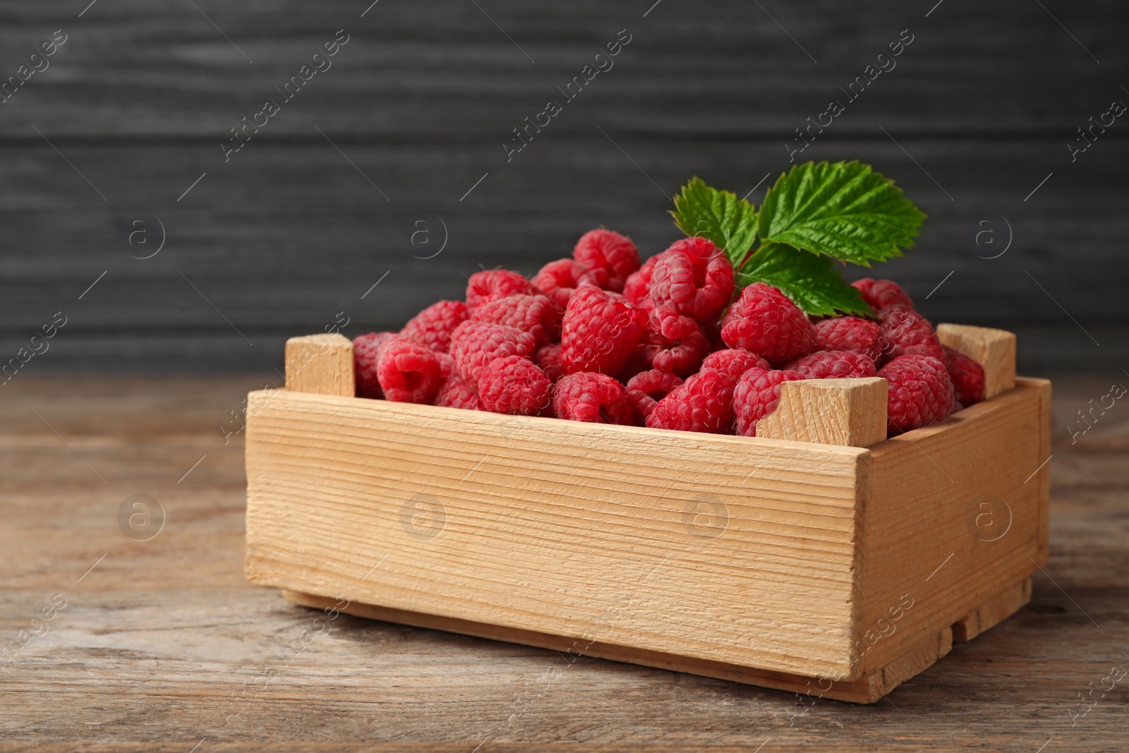 Photo of Crate of delicious fresh ripe raspberries with leaves on wooden table against dark background, space for text