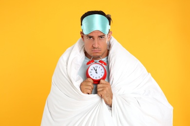 Man in sleeping mask wrapped with blanket holding alarm clock on yellow background