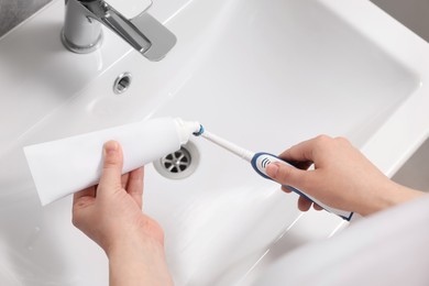 Photo of Woman squeezing toothpaste from tube onto toothbrush above sink in bathroom, closeup