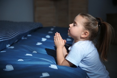 Photo of Little girl saying bedtime prayer near bed in room at night