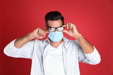 Photo of Man wiping foggy glasses caused by wearing medical mask on red background