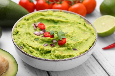 Photo of Bowl of delicious guacamole and ingredients on white wooden table, closeup