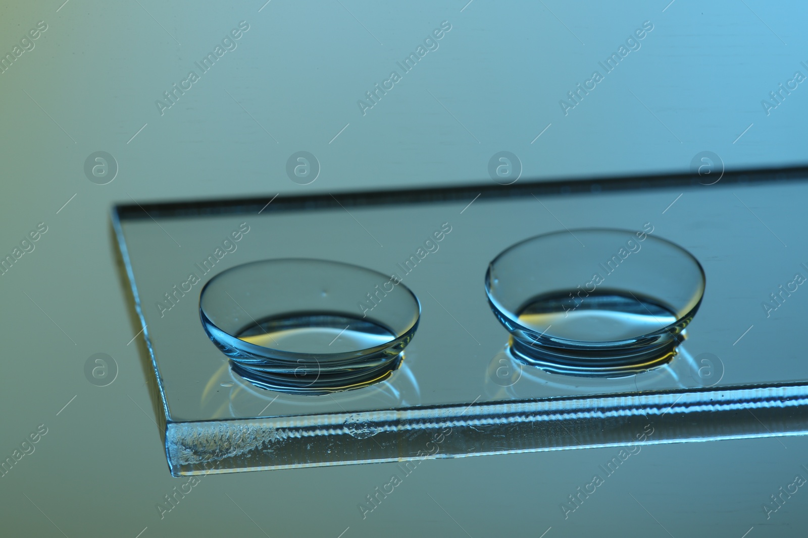Photo of Pair of contact lenses on glass against light blue background, closeup