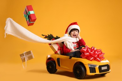 Photo of Cute little boy in Santa hat with Christmas tree and gift boxes driving children's electric toy car on yellow background