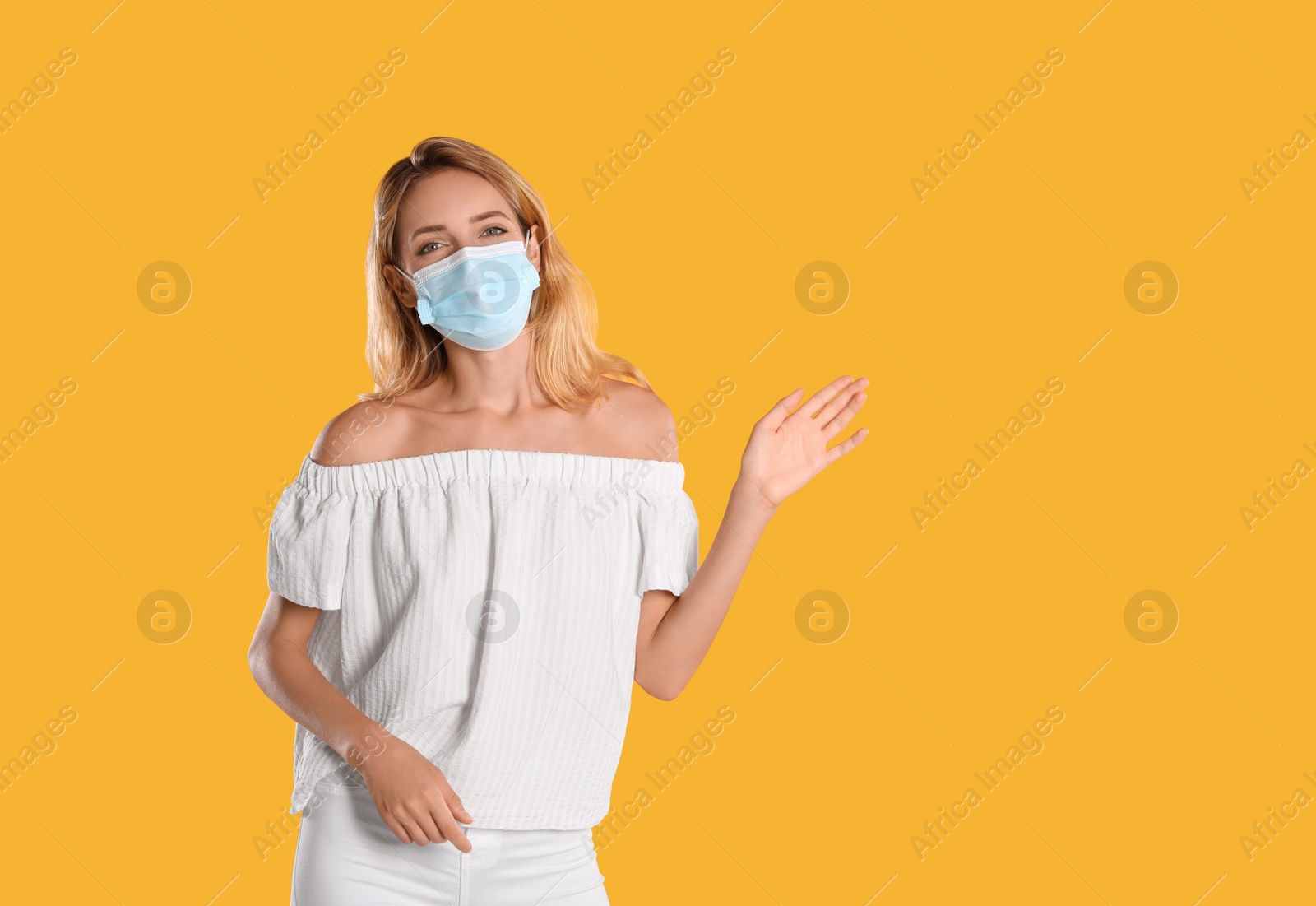 Photo of Woman in protective face mask showing hello gesture on yellow background, space for text. Keeping social distance during coronavirus pandemic