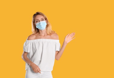 Photo of Woman in protective face mask showing hello gesture on yellow background, space for text. Keeping social distance during coronavirus pandemic