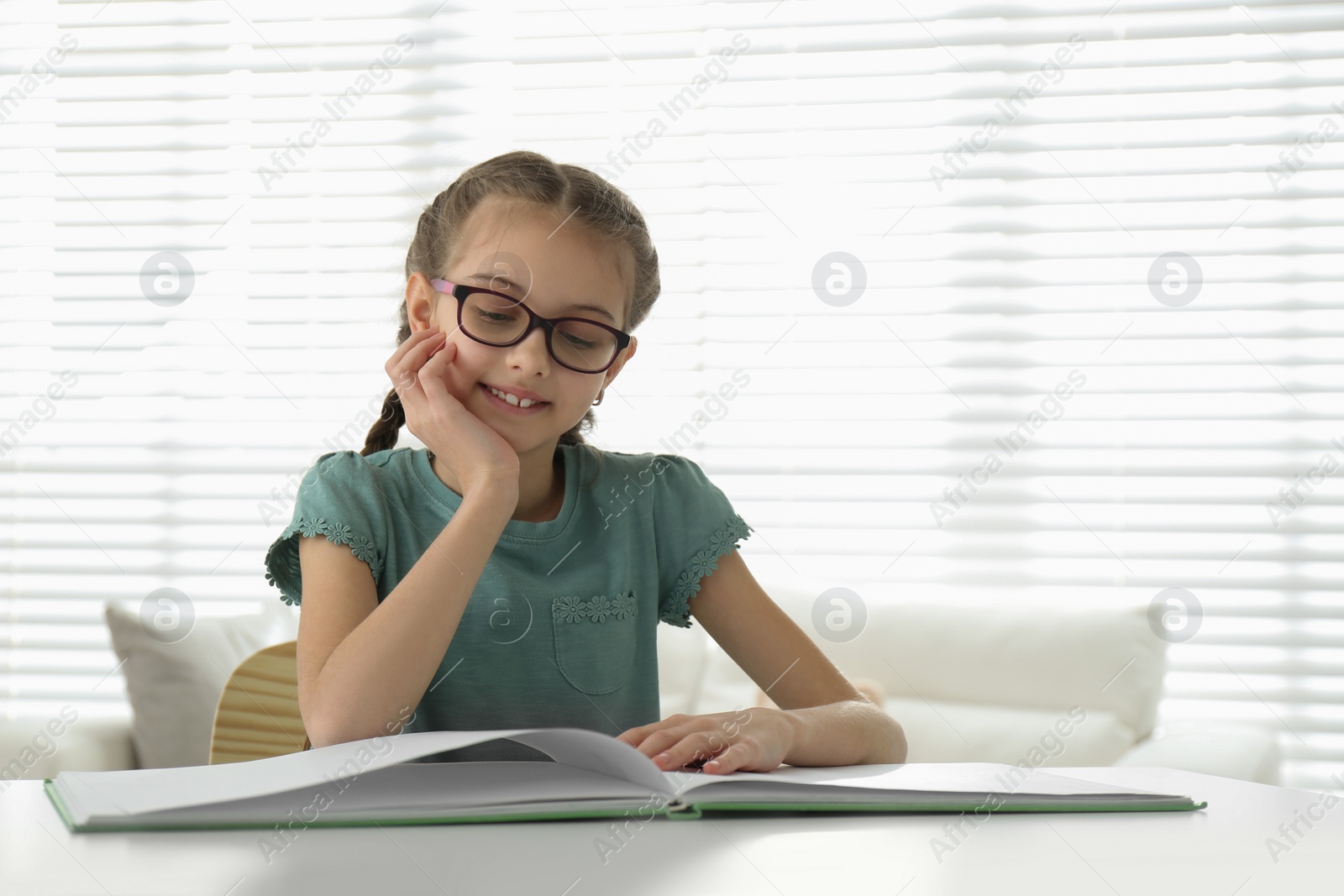 Photo of Cute little girl reading book at desk in room. Space for text