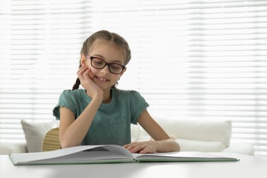 Photo of Cute little girl reading book at desk in room. Space for text