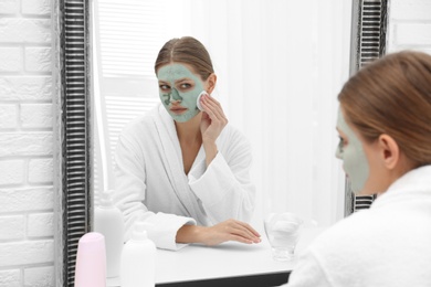 Photo of Beautiful woman removing homemade clay mask from her face at mirror indoors