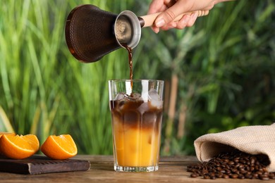 Photo of Woman pouring coffee into glass with orange juice and ice cubes at wooden table, closeup