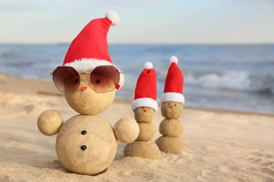 Snowmen made of sand with Santa hats and sunglasses on beach near sea. Christmas vacation