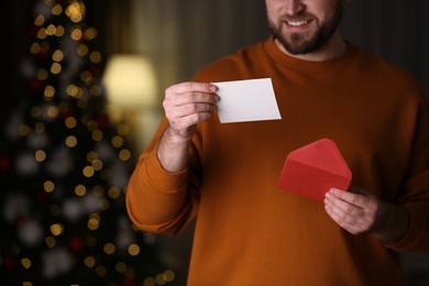 Man holding envelope and greeting card against blurred Christmas lights, closeup